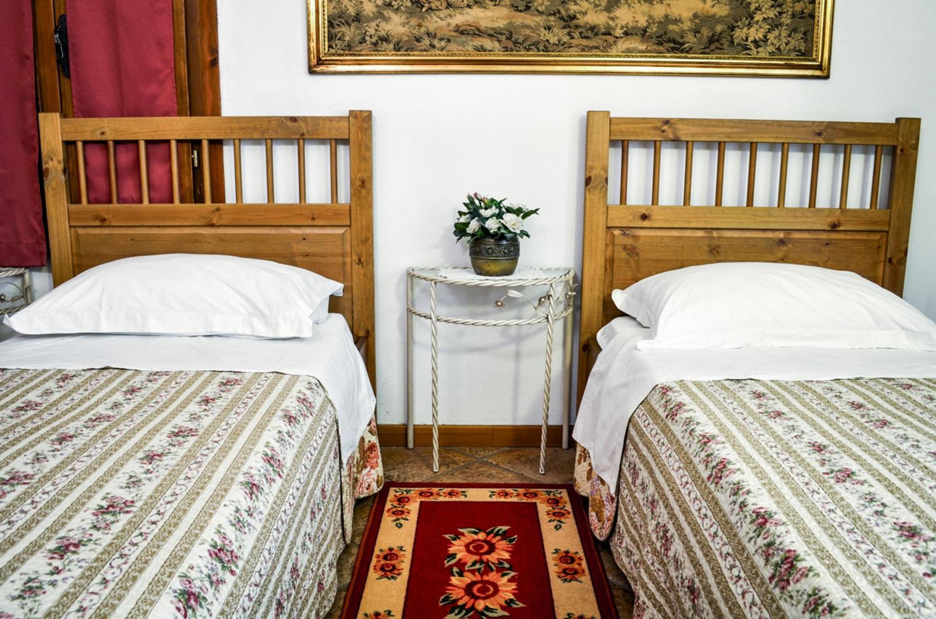 Una habitación con dos camas de madera, ropa de cama blanca, una mesita redonda con una planta y un tapete floral rojo.
