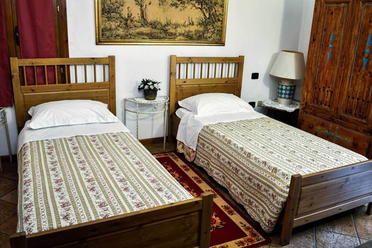 Een kamer met twee houten bedden, elk voorzien van een wit kussen en een bedovertrek met bloemenprint. Er staat een klein rond tafeltje met een lamp en een potplant tussen de bedden.