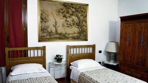 A room with two single beds, a wooden headboard, a small round table with a lamp, a large framed painting on the wall, and a wooden wardrobe.