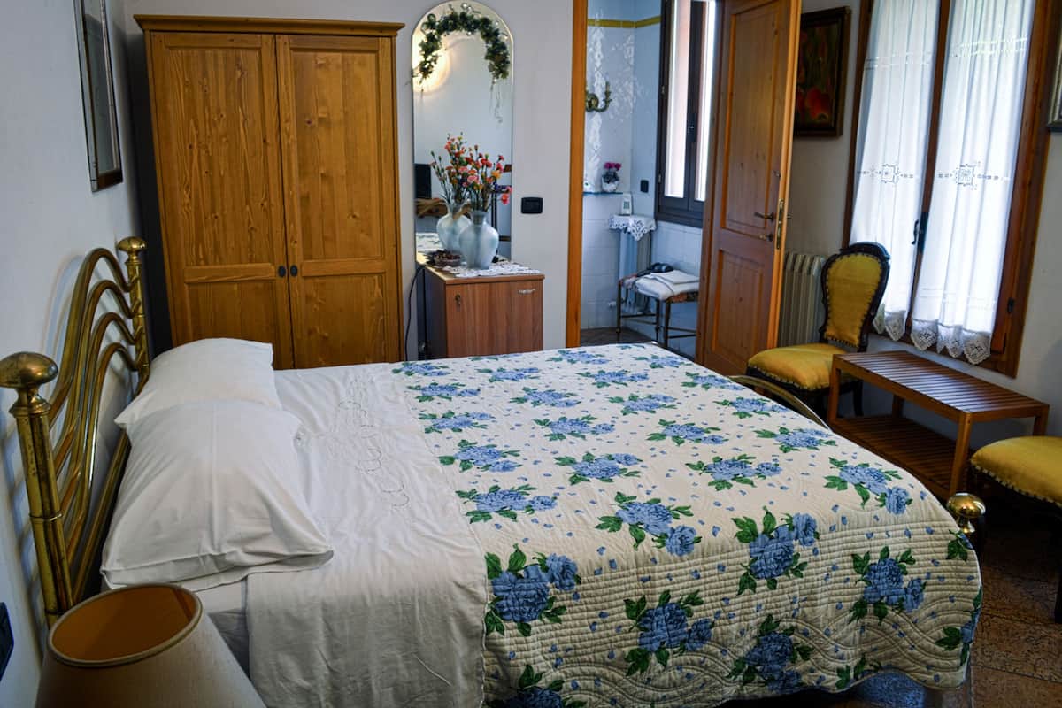 Een slaapkamer met een bed, een houten garderobe, een klein bureau met een bloevas, een stoel en een raam met gordijnen.