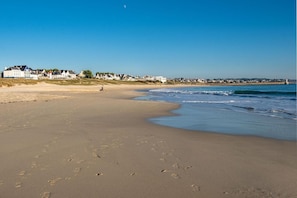 Uma praia com pegadas que levam à água, céu claro e casas ao fundo.