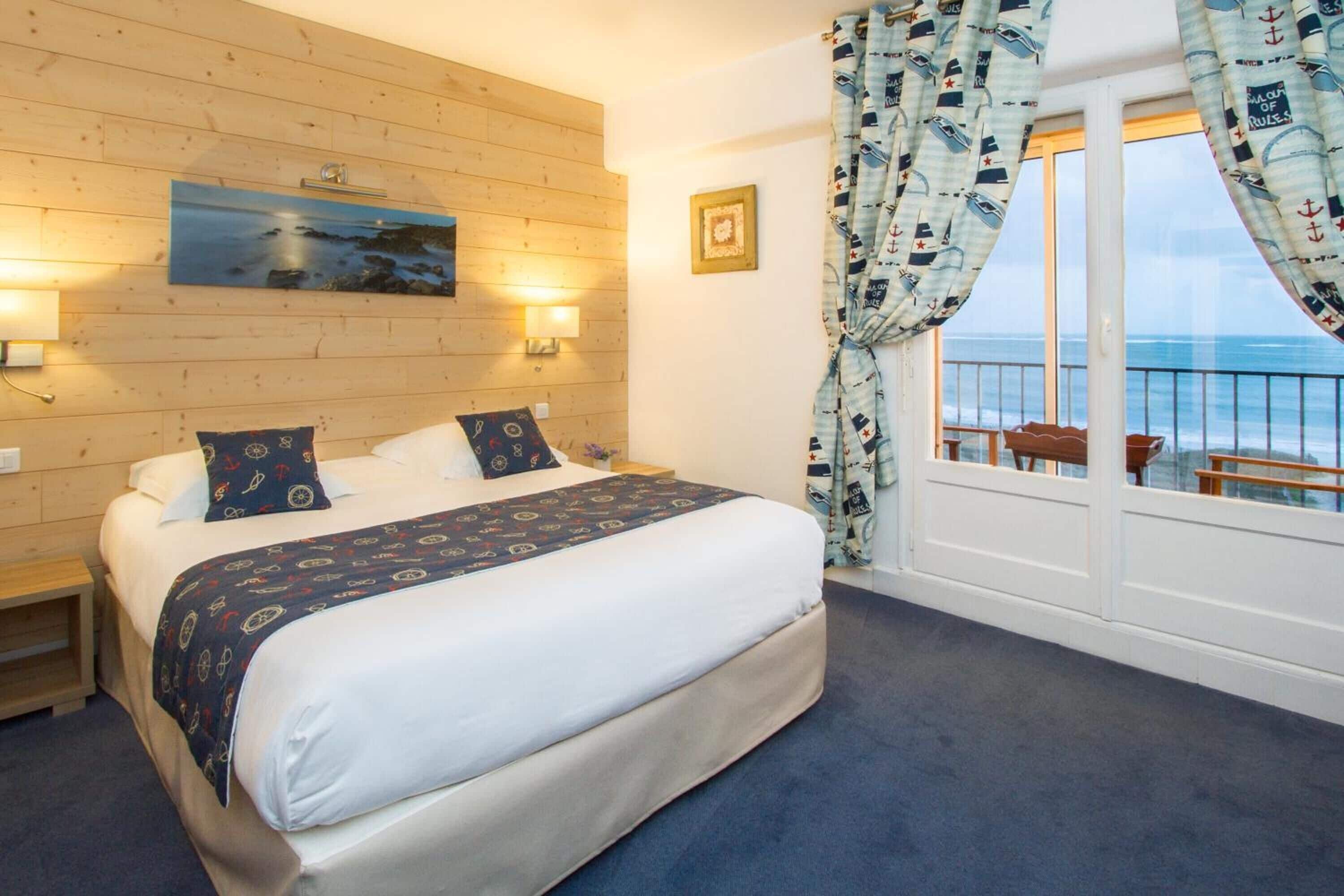 A bedroom with a large bed, a wooden wall, a painting, and a view of the sea through a window with nautical curtains.