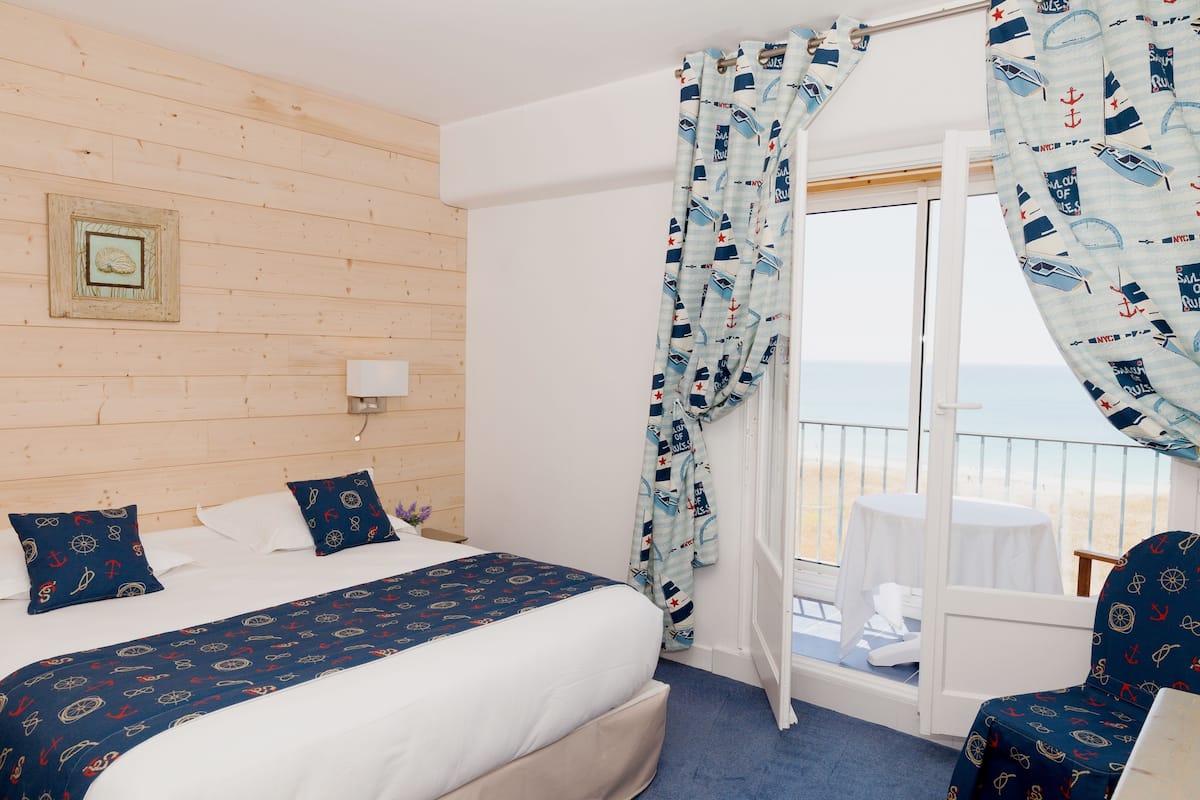Ein Hotelzimmer mit Bett, Sessel, Fenster mit Vorhängen und Blick auf den Strand.