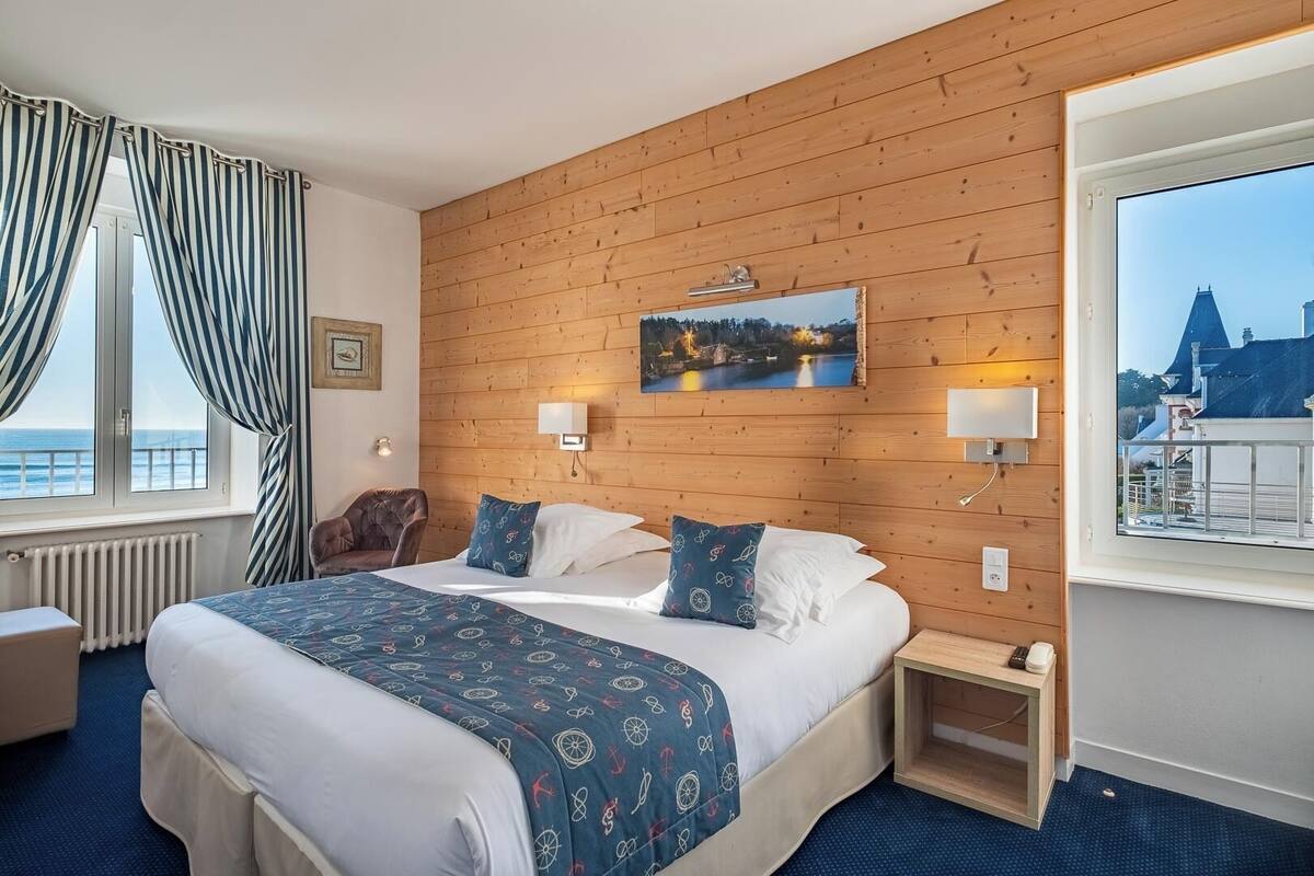 Ein Hotelzimmer mit einer Holzwand, einem großen Bett mit gemusterten Betttextilien, Meerblick durch ein Fenster und einem kleinen Nachttisch mit Lampe.