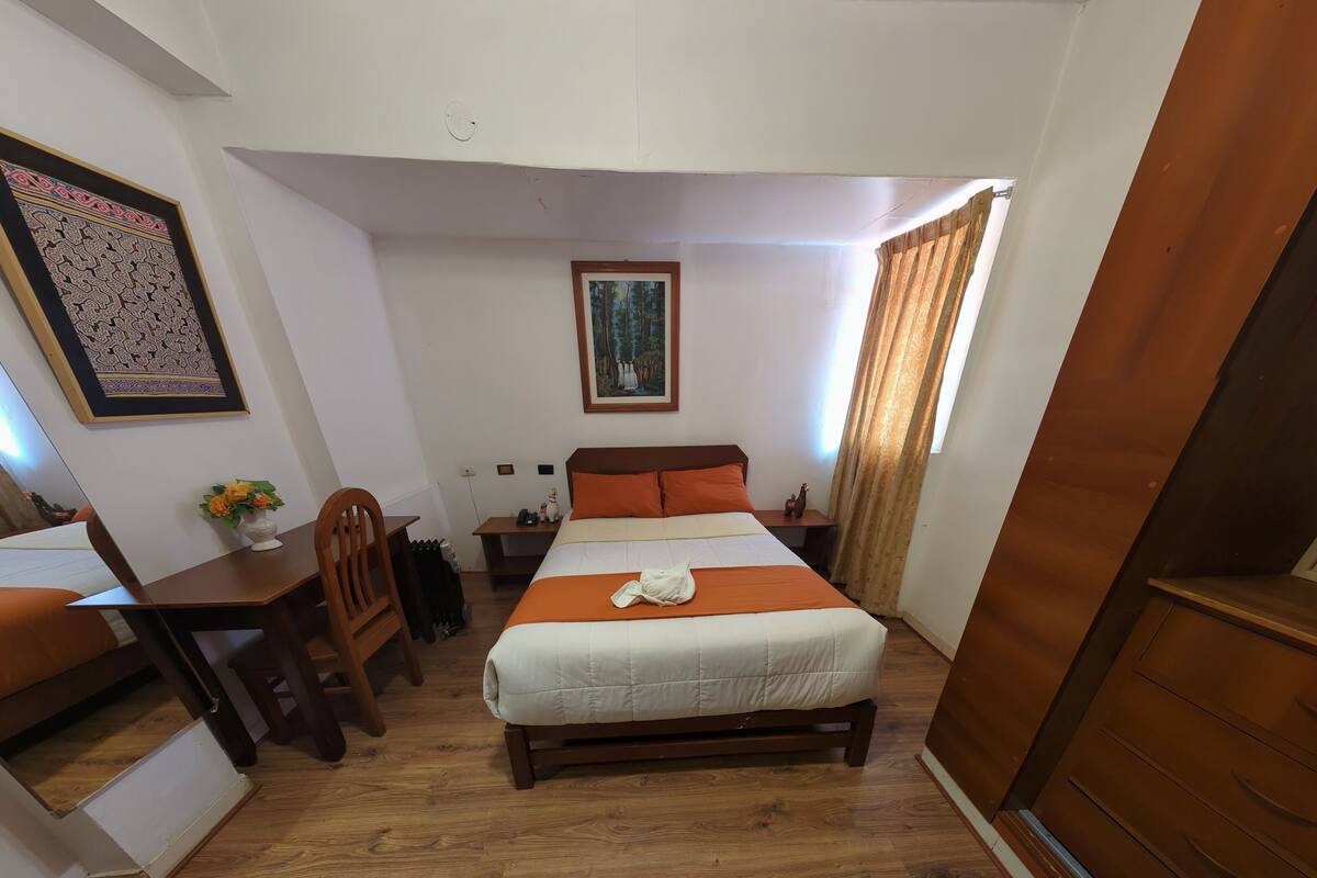 A bedroom with a bed, wooden floor, a desk with a chair, a framed picture, and a window with curtains.