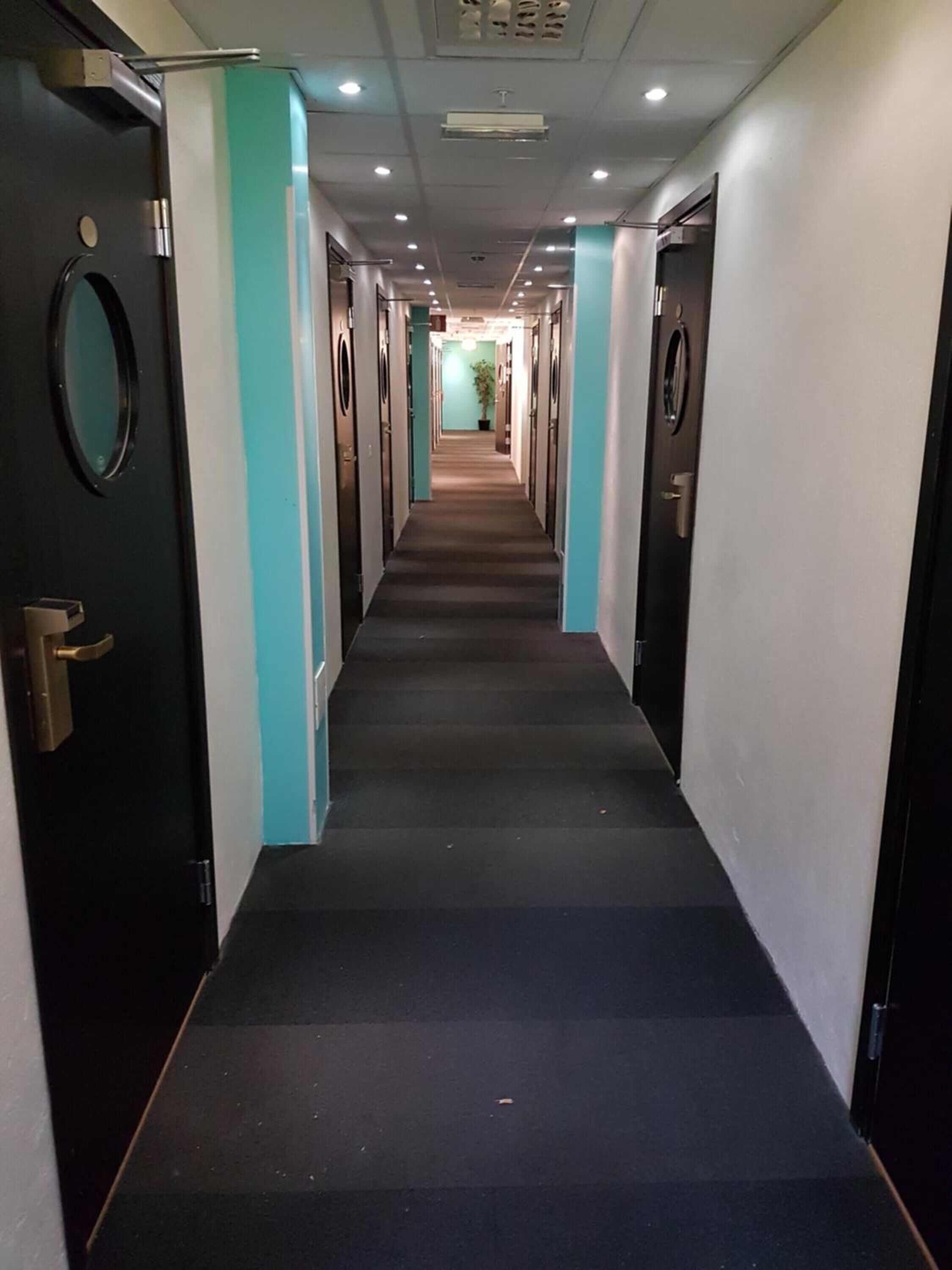 Un largo pasillo de hotel, estrecho, con puertas a ambos lados, suelo alfombrado e iluminación empotrada en el techo.