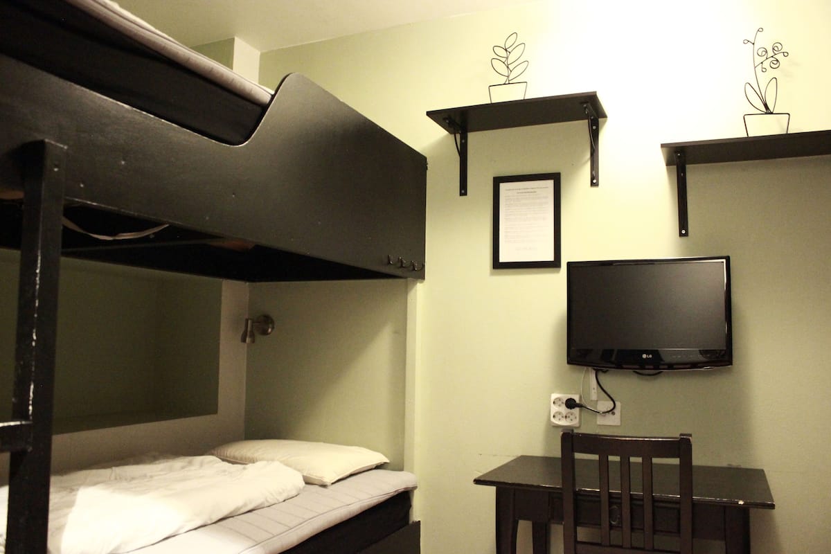 Una cama litera con escritorio y silla, un televisor y dos estantes en la pared con objetos decorativos.