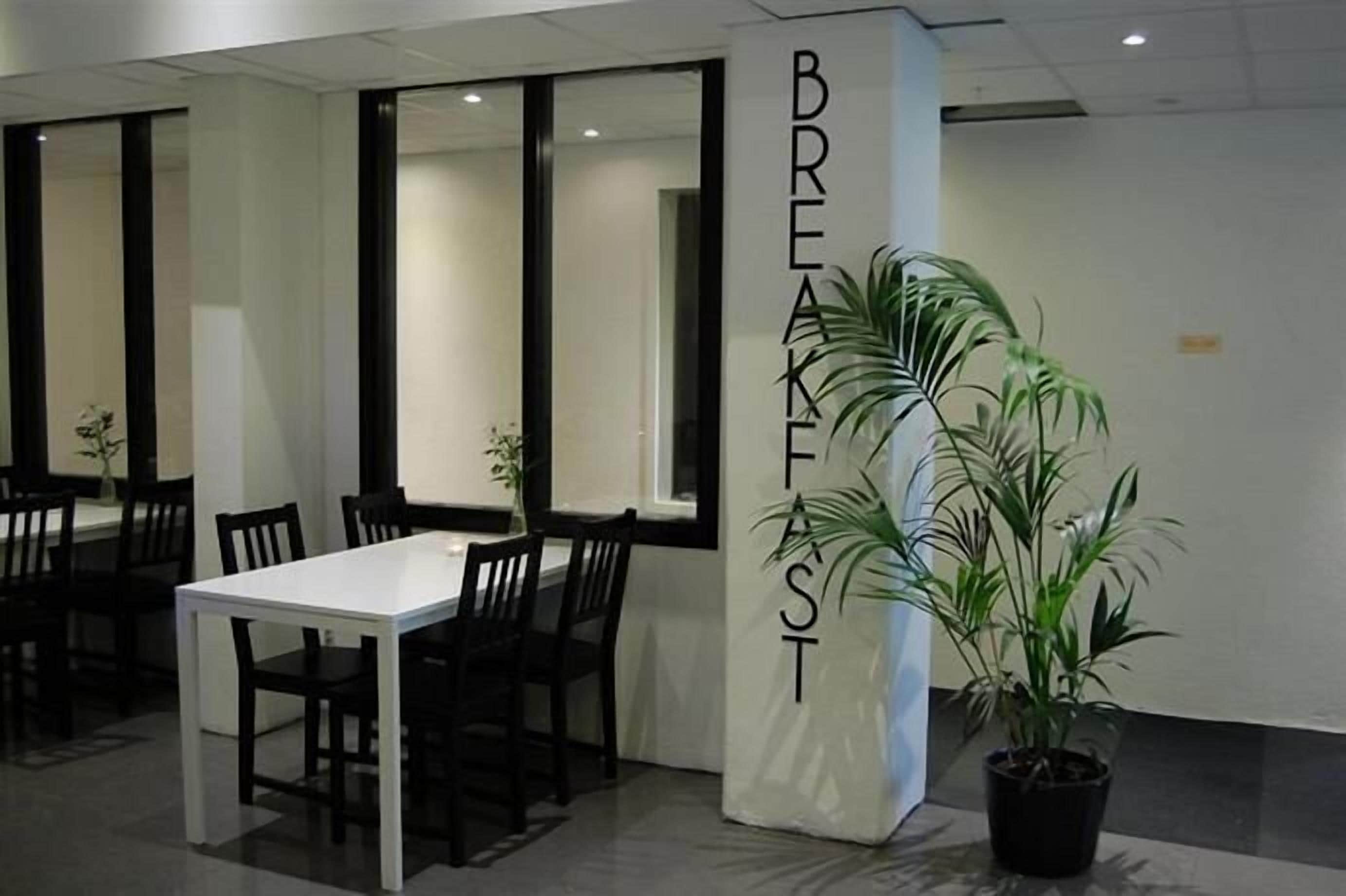 Un moderno comedor con una mesa blanca y sillas negras, una planta grande en maceta y un pilar con la palabra "BREAKFAST".