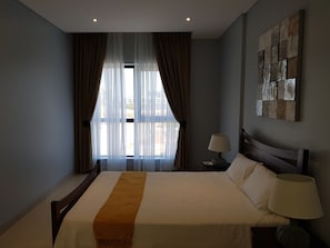 Habitación de hotel con una cama grande, una mesita de noche con lámpara, una ventana con cortinas y una obra de arte colgada en la pared.