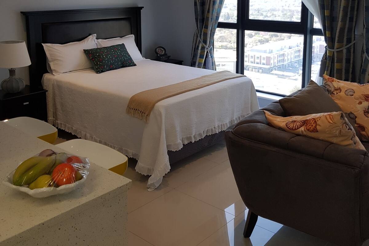 A hotel room with a bed, a chair, a small table with fruit, and a view through large windows.