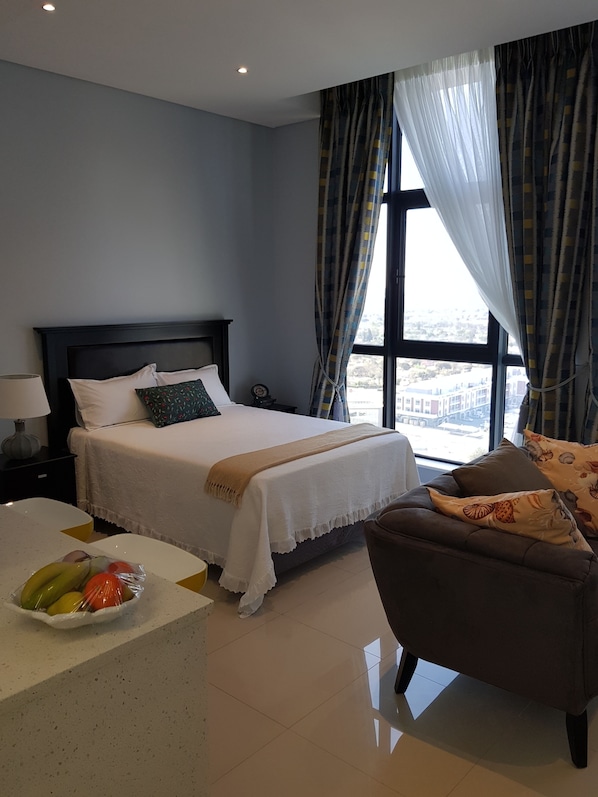 Habitación de hotel con cama, una silla, una mesita con fruta y vistas amplias a través de grandes ventanales.