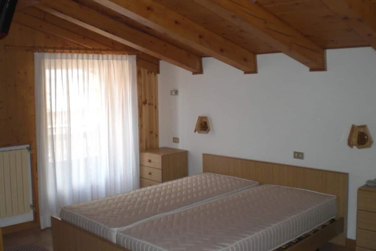 A room with a wooden ceiling, a bed with a mattress, a wooden dresser, and a window with curtains.