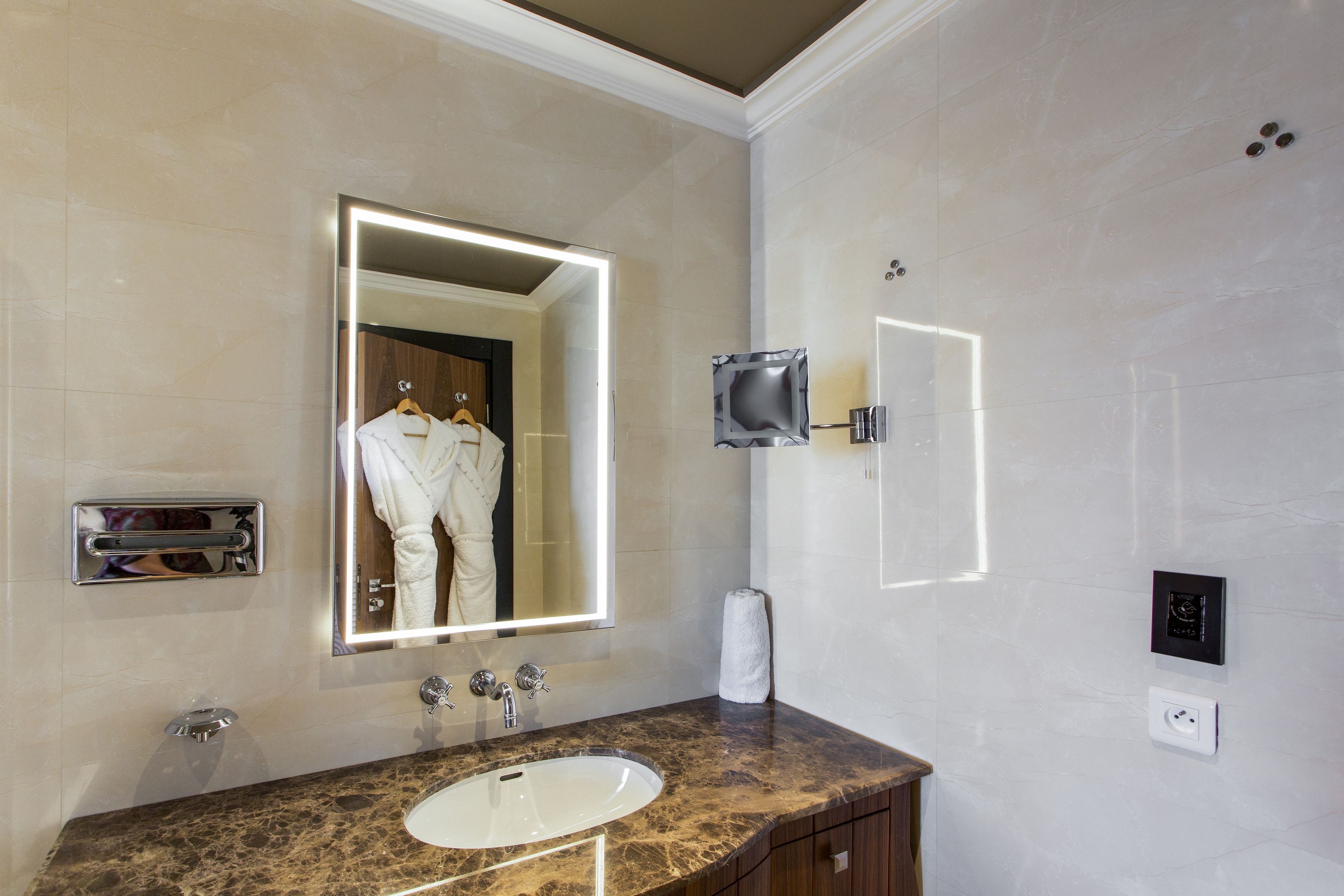Une salle de bain dotée d’un lavabo en marbre, d’un miroir avec éclairage intégré, d’un distributeur de serviettes et d’un thermostat fixé au mur.