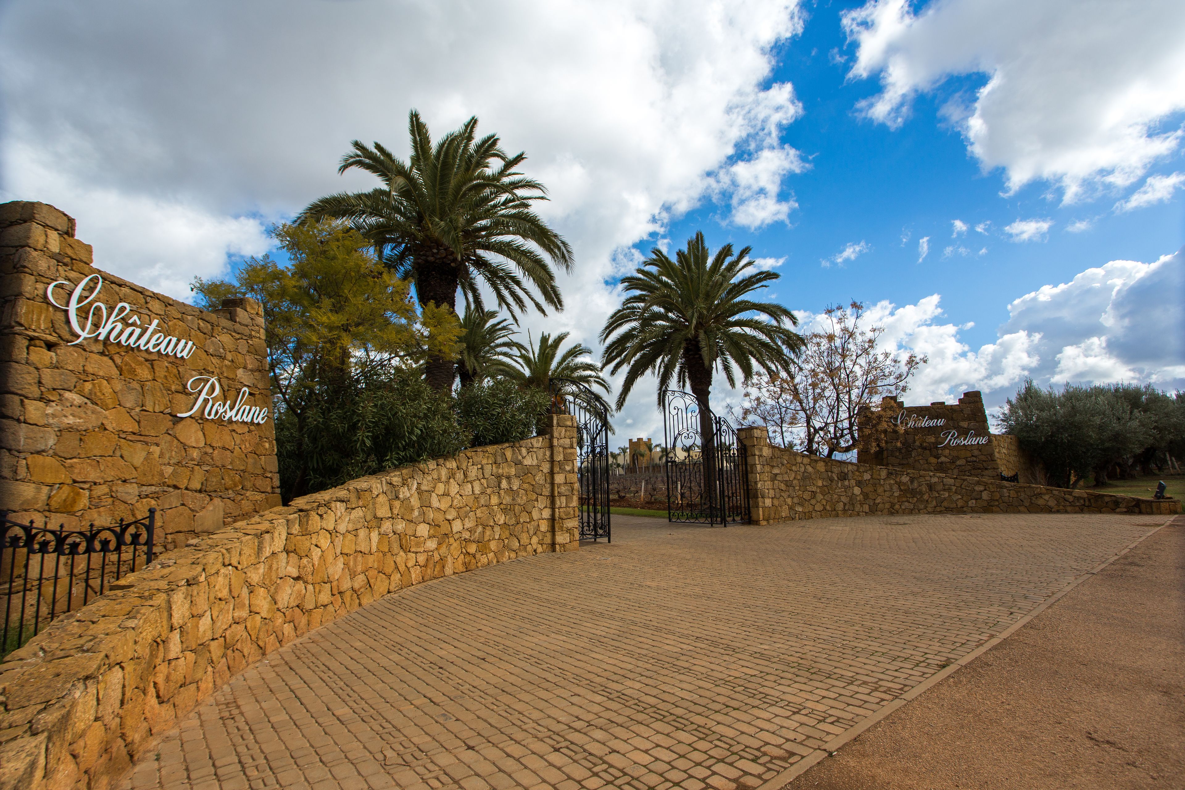 Um muro de pedra com uma placa indicando "Chateau Roslane" e um caminho pavimentado que leva a um portão.
