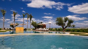 Una gran piscina al aire libre con sillones de descanso y sombrillas, rodeada de palmeras y un cielo azul despejado.