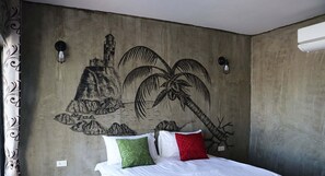 Un dormitorio con un mural de una montaña y palmeras, una cama con ropa de cama blanca y dos almohadas, y un aire acondicionado de pared.