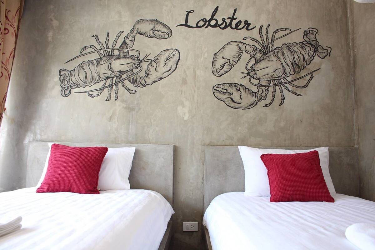 A hotel room with two beds, each with white bedding and red pillows. The walls feature a large drawing of lobsters and the word "Lobster" above it.