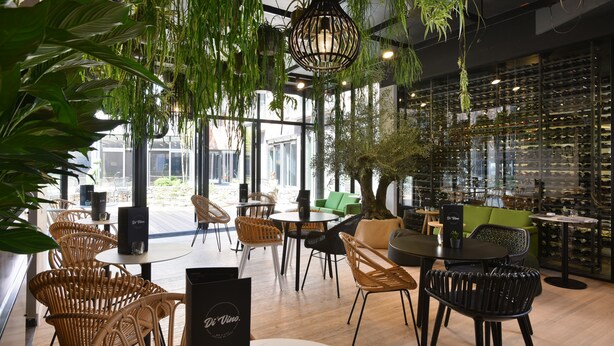 Un restaurant avec des chaises et des tables en osier, des plantes suspendues et un présentoir à vin en arrière-plan.