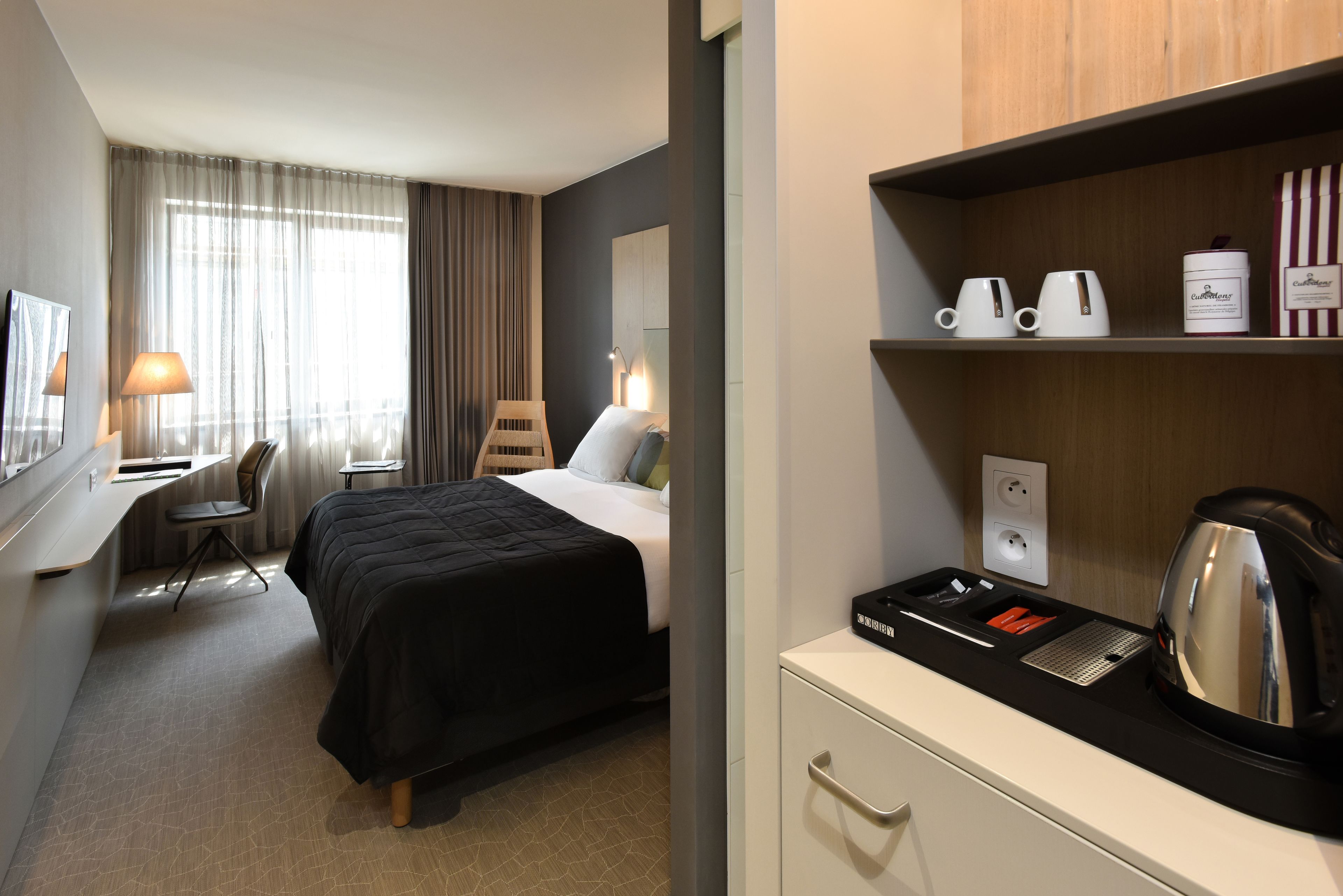 A hotel room with a bed, desk, chair, and a kitchenette area with a kettle and coffee cups.