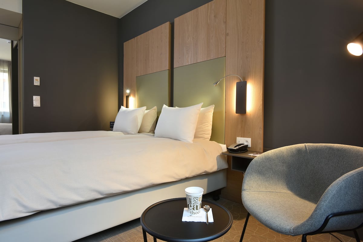 Una habitación de hotel con una cama grande, un sillón gris y una mesita con una taza y una cuchara.
