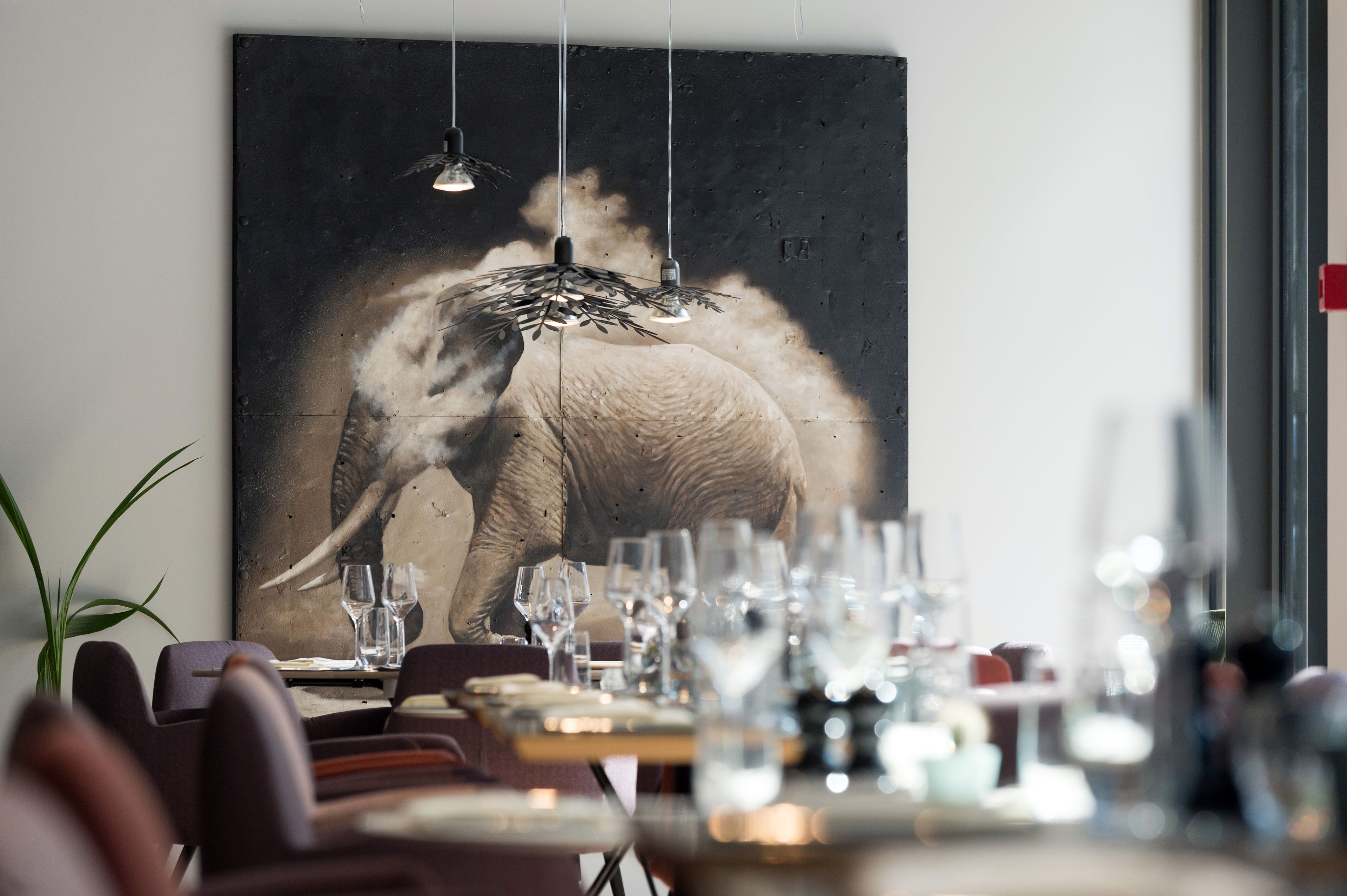A dining area with a large elephant painting on the wall, a table set with glasses and plates, and hanging pendant lights.