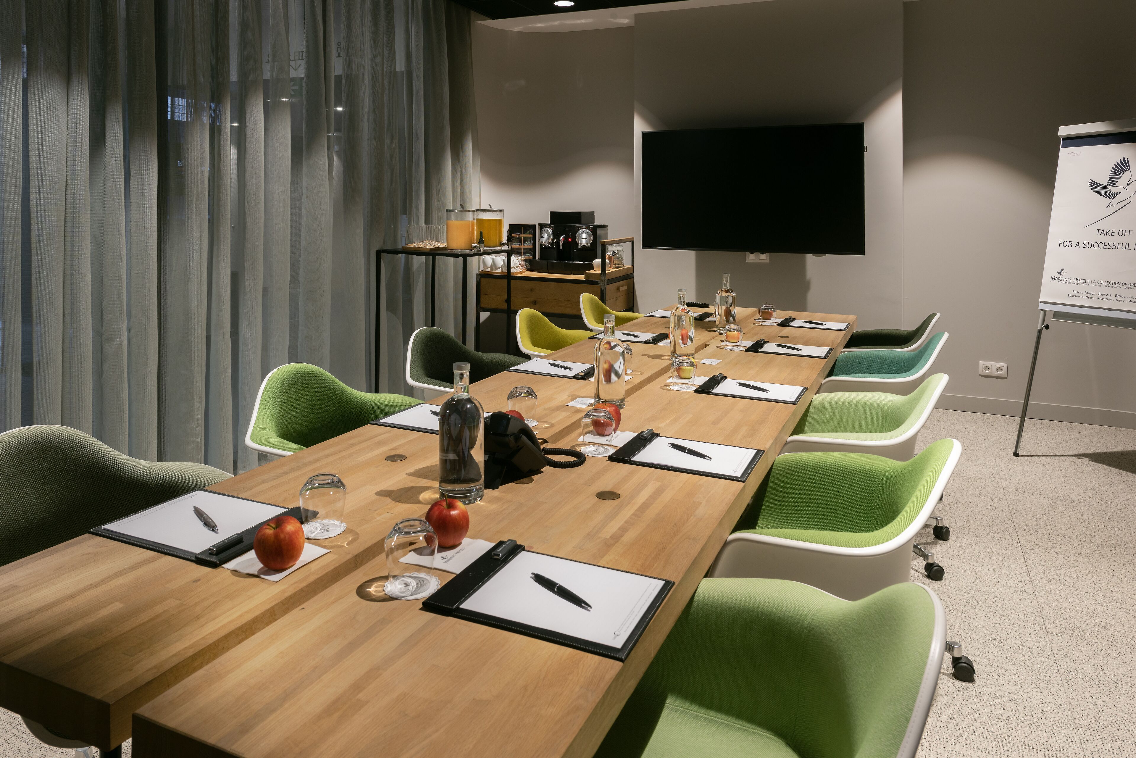 Ein Konferenzraum mit langem Tisch, grünen Stühlen, einer Projektionsleinwand und einer Whiteboard.