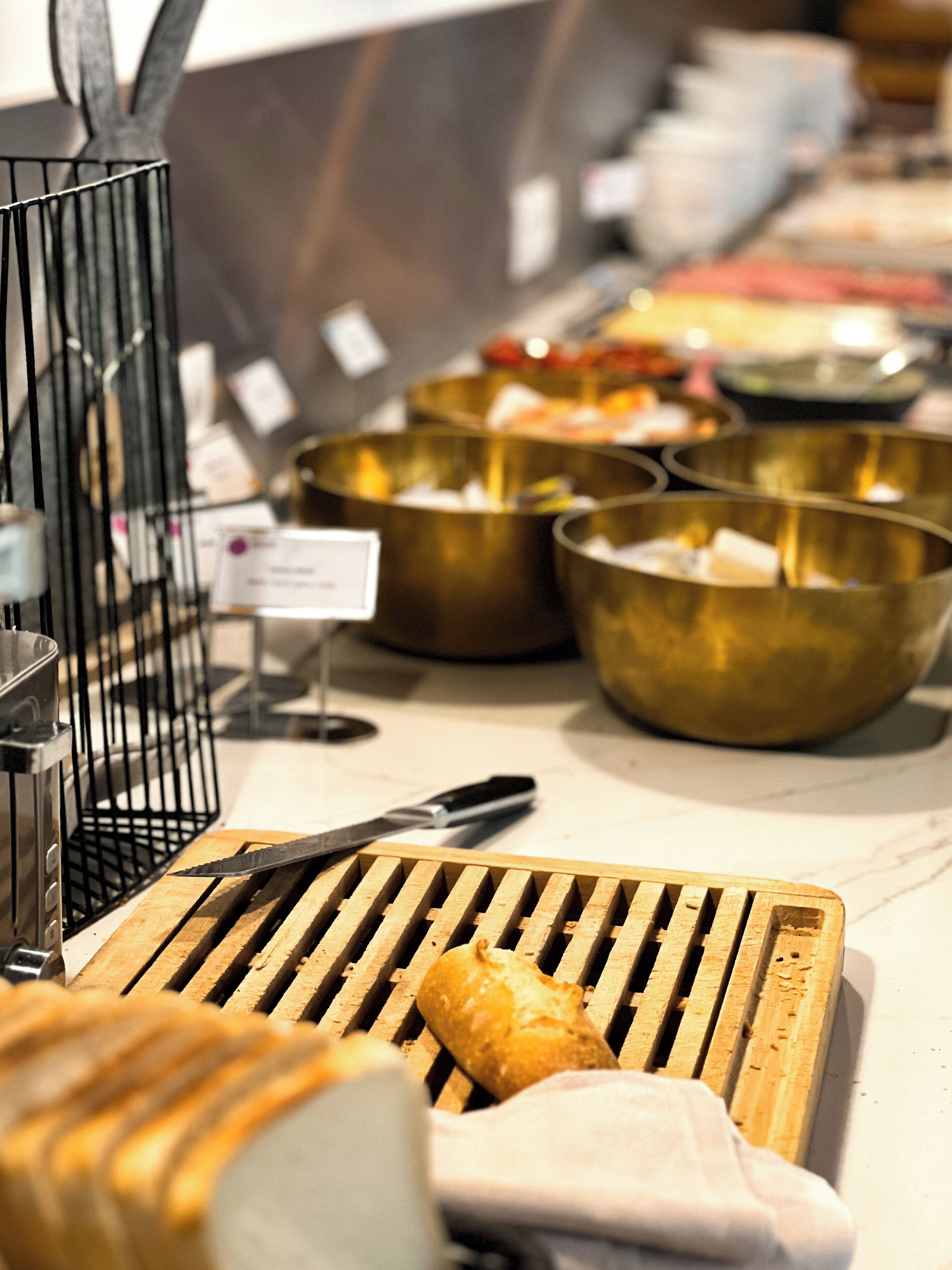 Montaje de bufé con diferentes alimentos en recipientes metálicos, un cortador de pan y un cuchillo sobre una tabla de cortar.