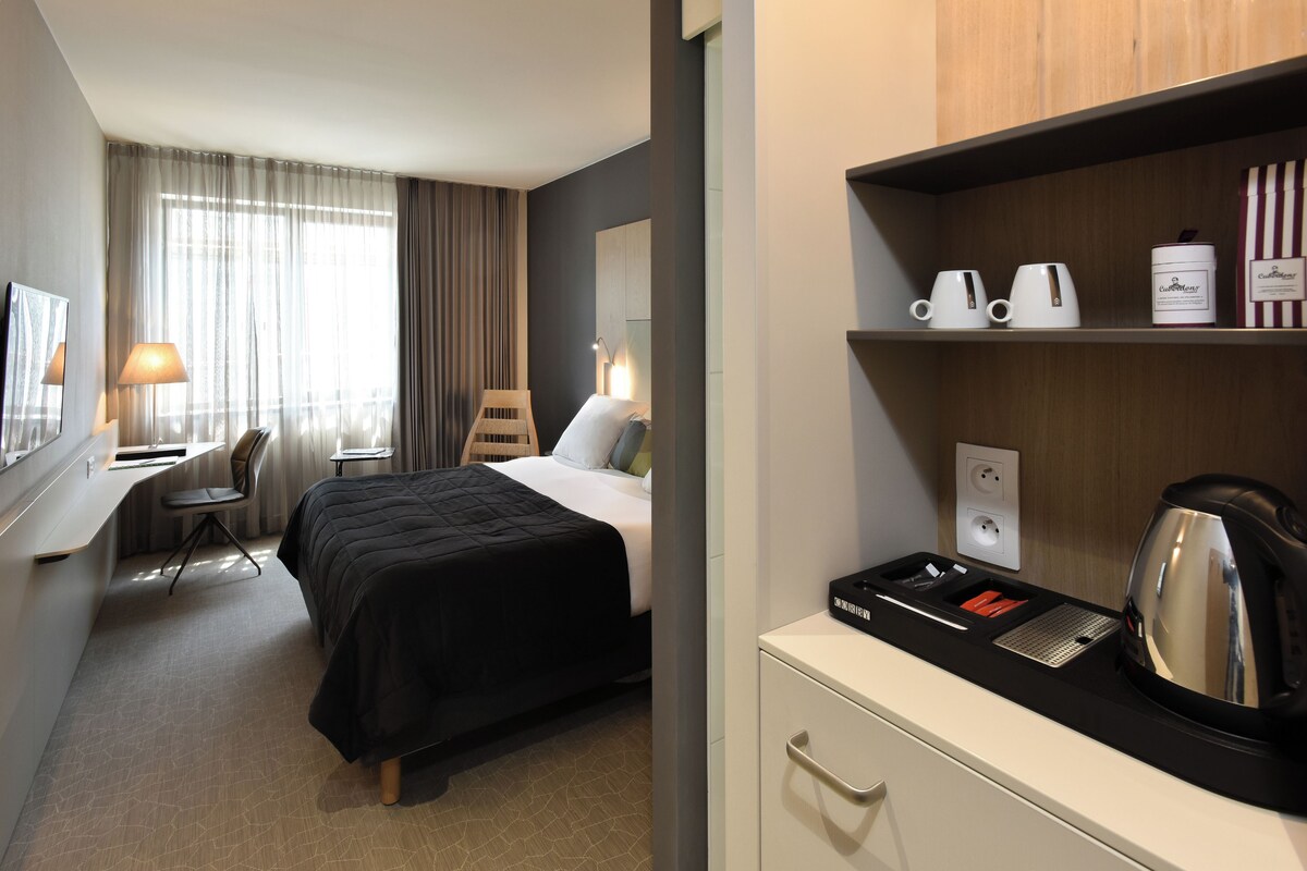 A hotel room with a bed, desk, chair, and a kitchenette area with a kettle and coffee cups.