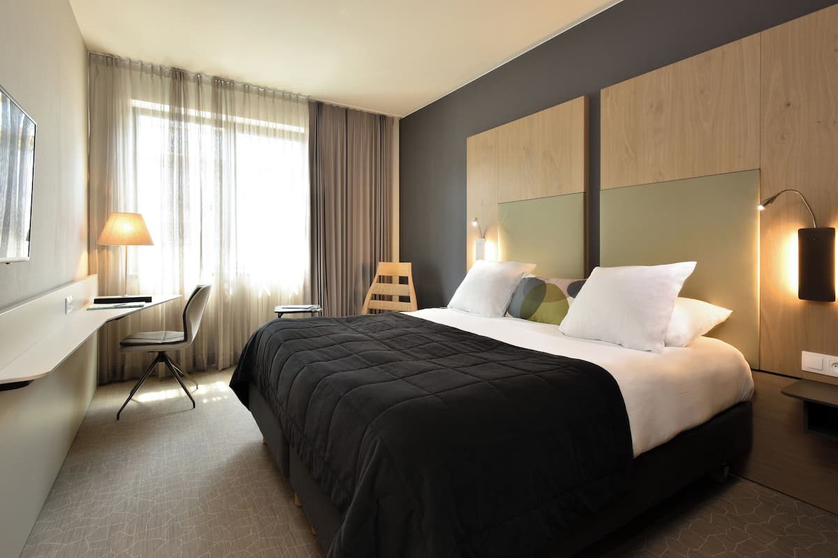Habitación de hotel con cama, escritorio, silla y una ventana grande con cortinas.