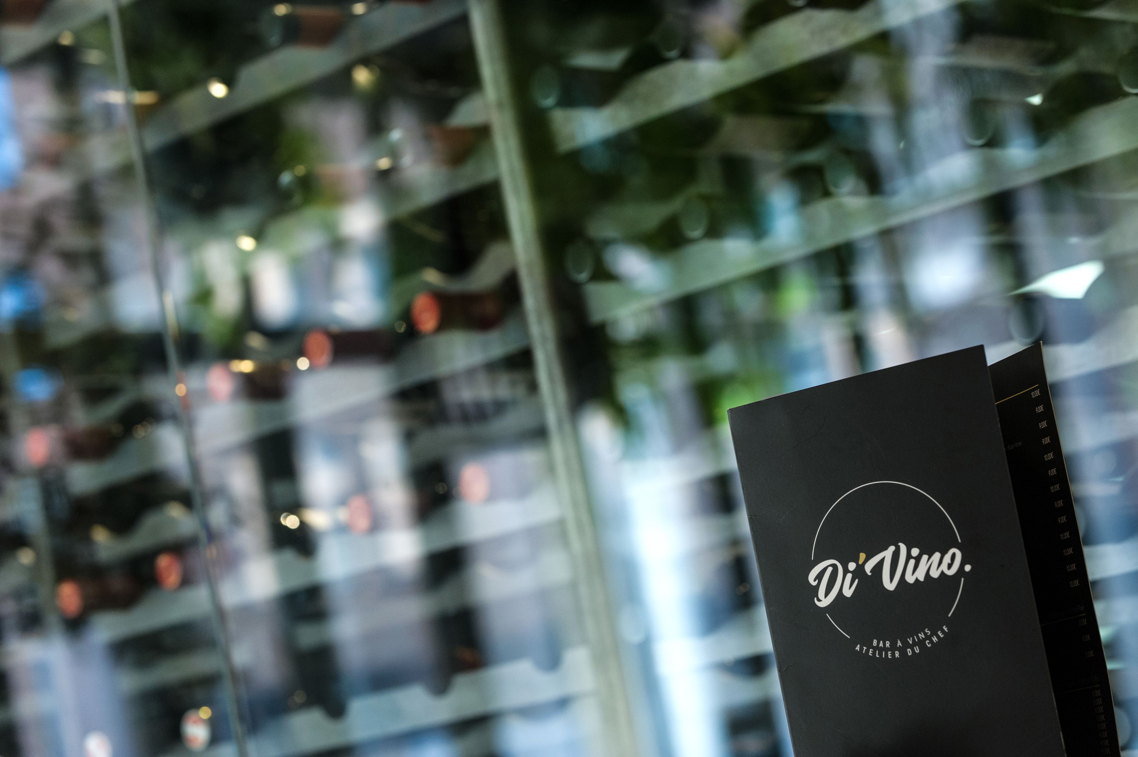 A black brochure with the text "Di Vino." and "BAR A VINS, ATELIER DU CHEF" on a glass window.