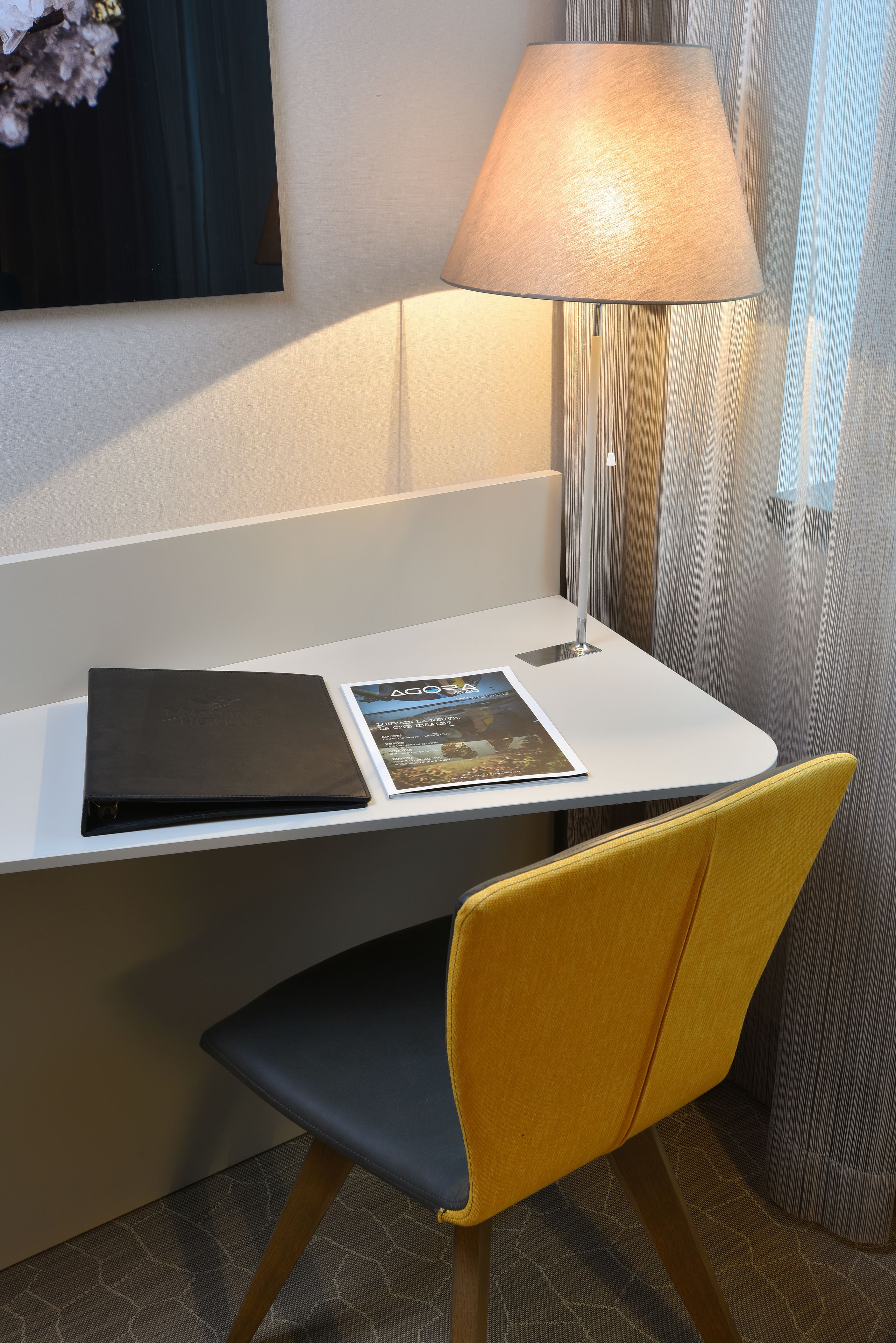 A hotel room with a desk, a lamp, a magazine, and a chair.