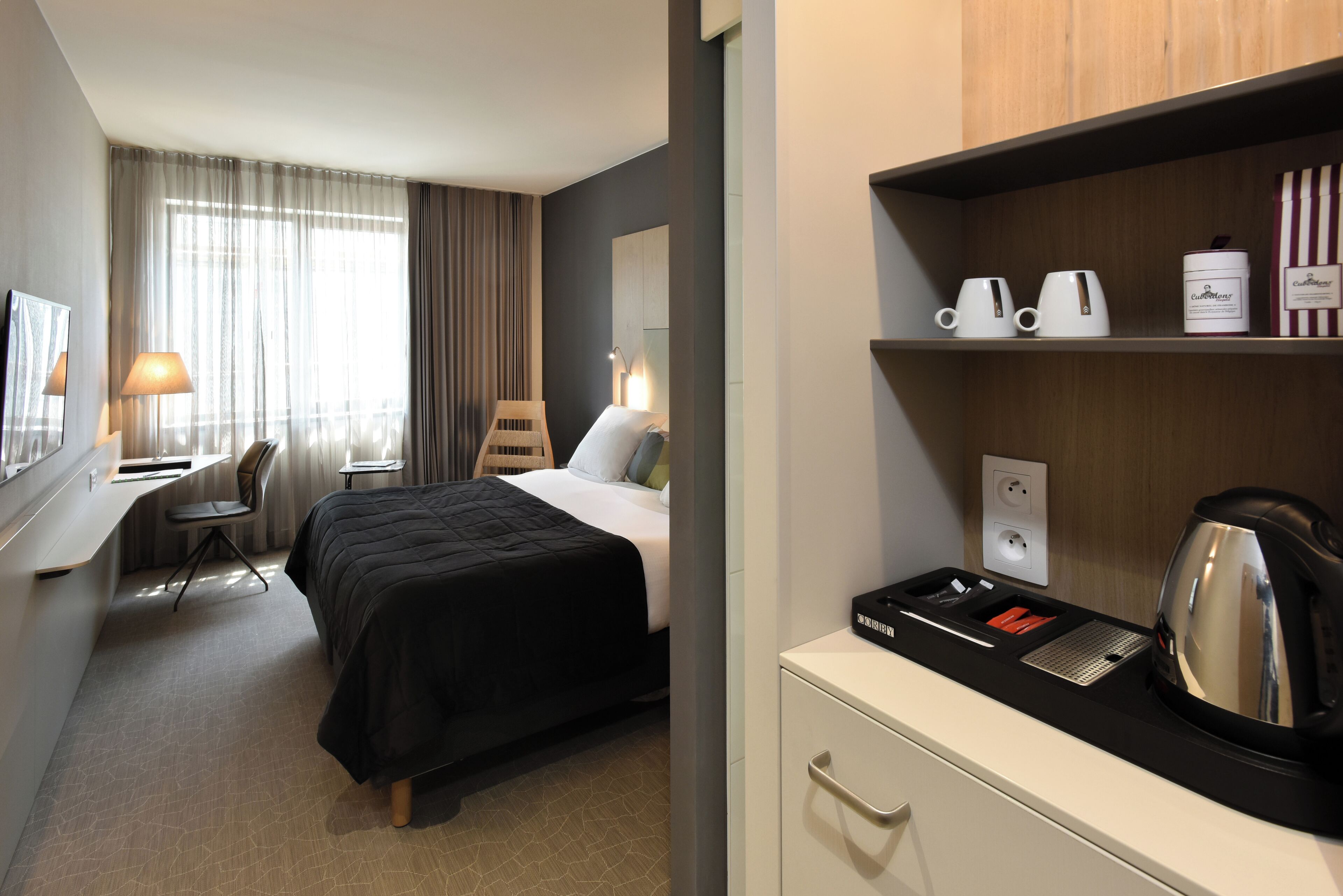 Una habitación de hotel con cama, escritorio, silla y una zona de kitchenette con hervidor y tazas de café.