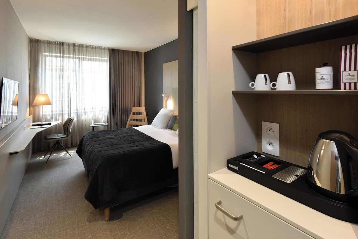 Una habitación de hotel con cama, escritorio, silla y una zona de kitchenette con hervidor y tazas de café.