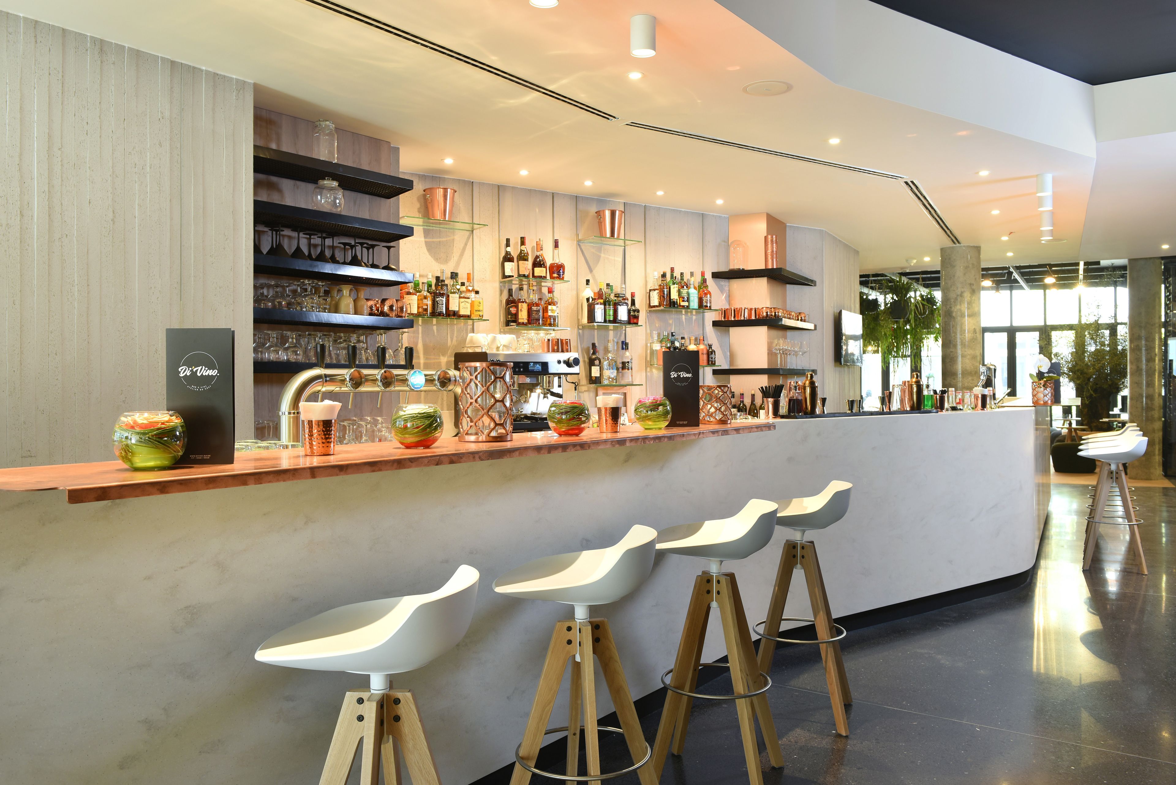 A modern bar area with a long counter, multiple shelves of bottles, and three white bar stools.