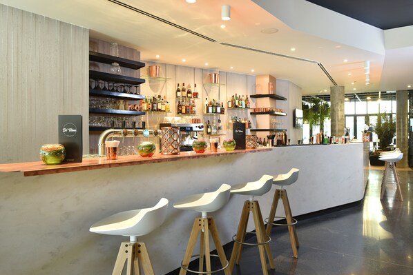 Zona de bar moderna con una barra larga, varias estanterías llenas de botellas y tres taburetes blancos.
