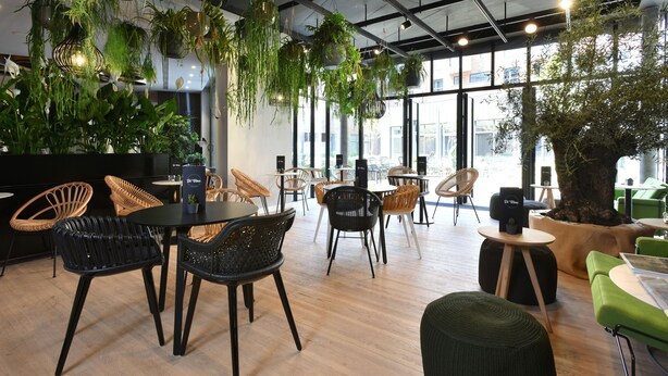 A modern indoor space with multiple seating areas, including round tables and chairs, surrounded by hanging plants and a large tree. - Martin's All Suites (Ottignies-Louvain-la-Neuve)