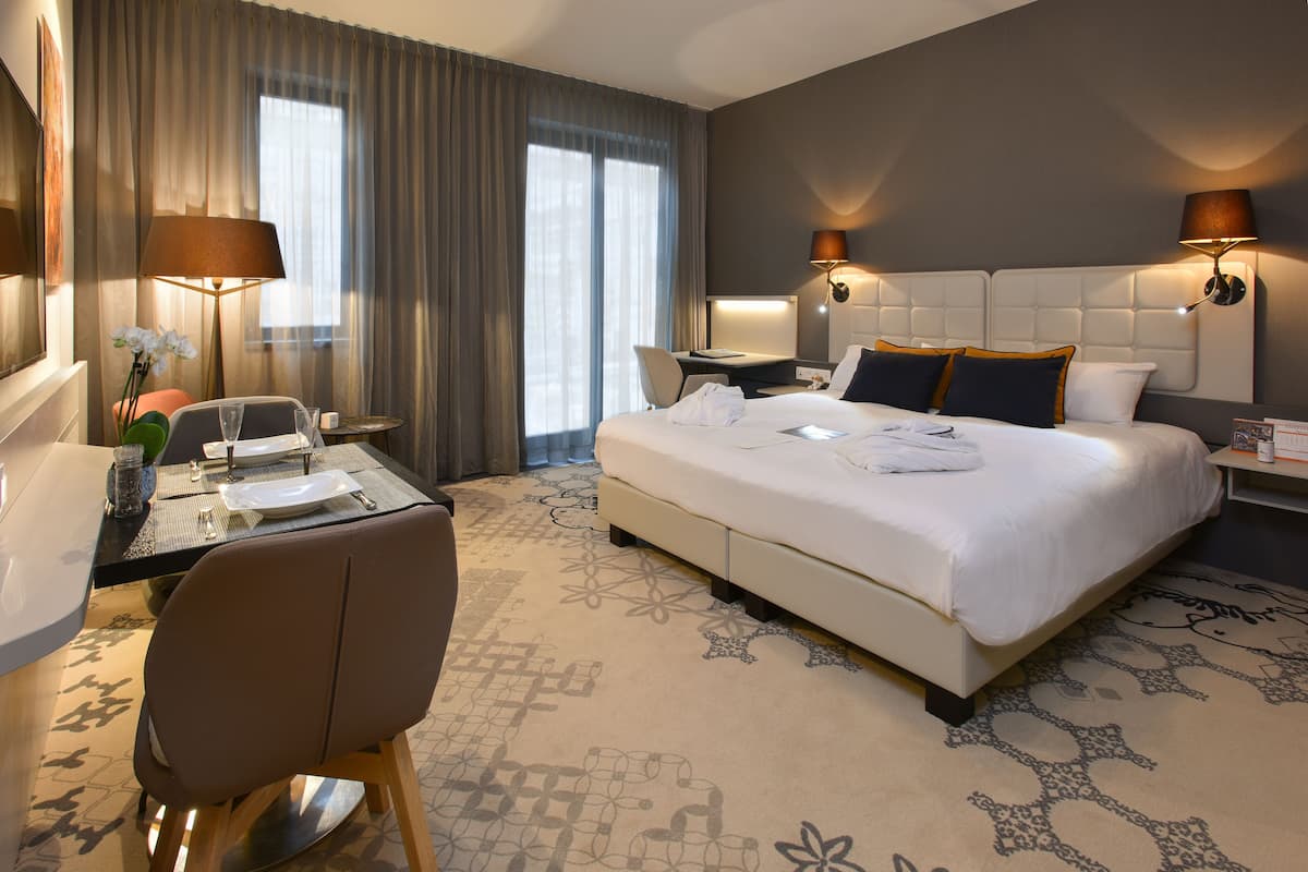A hotel room with a large bed, two bedside tables, a desk with a chair, a dining area, and a large window with curtains.