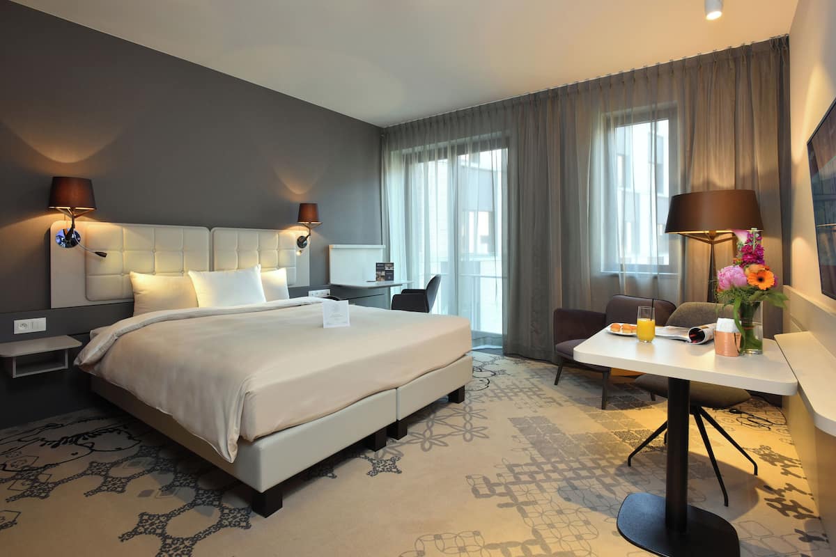 A hotel room with a large bed, bedside tables, a desk, a chair, a small table with a lamp and flowers, and a view of the outdoors through the window.