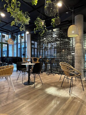 Un interior moderno con suelo de madera, sillas negras y de mimbre, una mesa, plantas colgantes y una estantería para vinos al fondo.