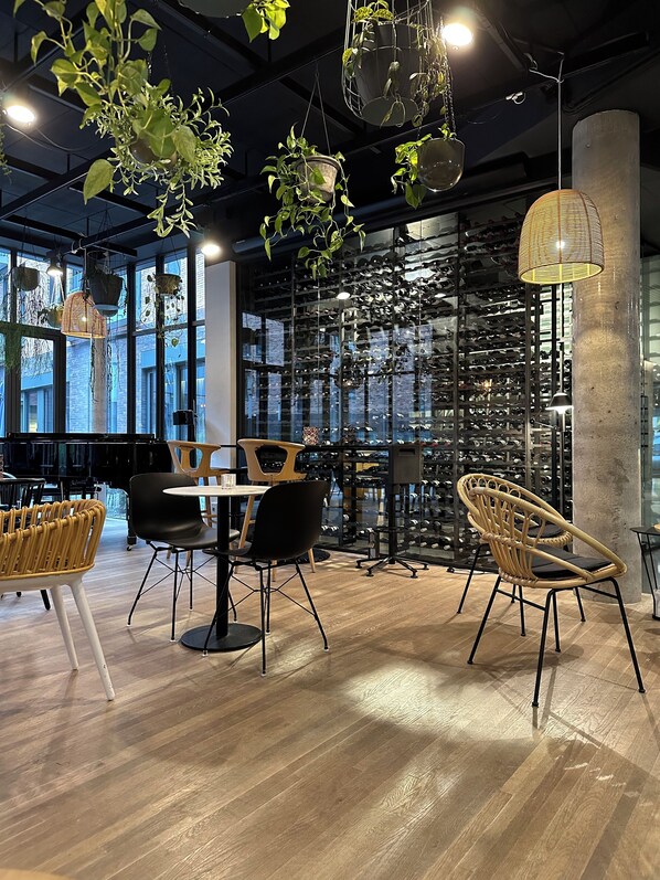 Un interior moderno con suelo de madera, sillas negras y de mimbre, una mesa, plantas colgantes y una estantería para vinos al fondo.