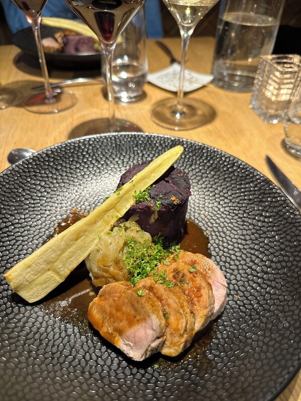 Un plato de comida con un trozo de carne oscura como centro, un pescado y algunas verduras sobre un plato con textura.