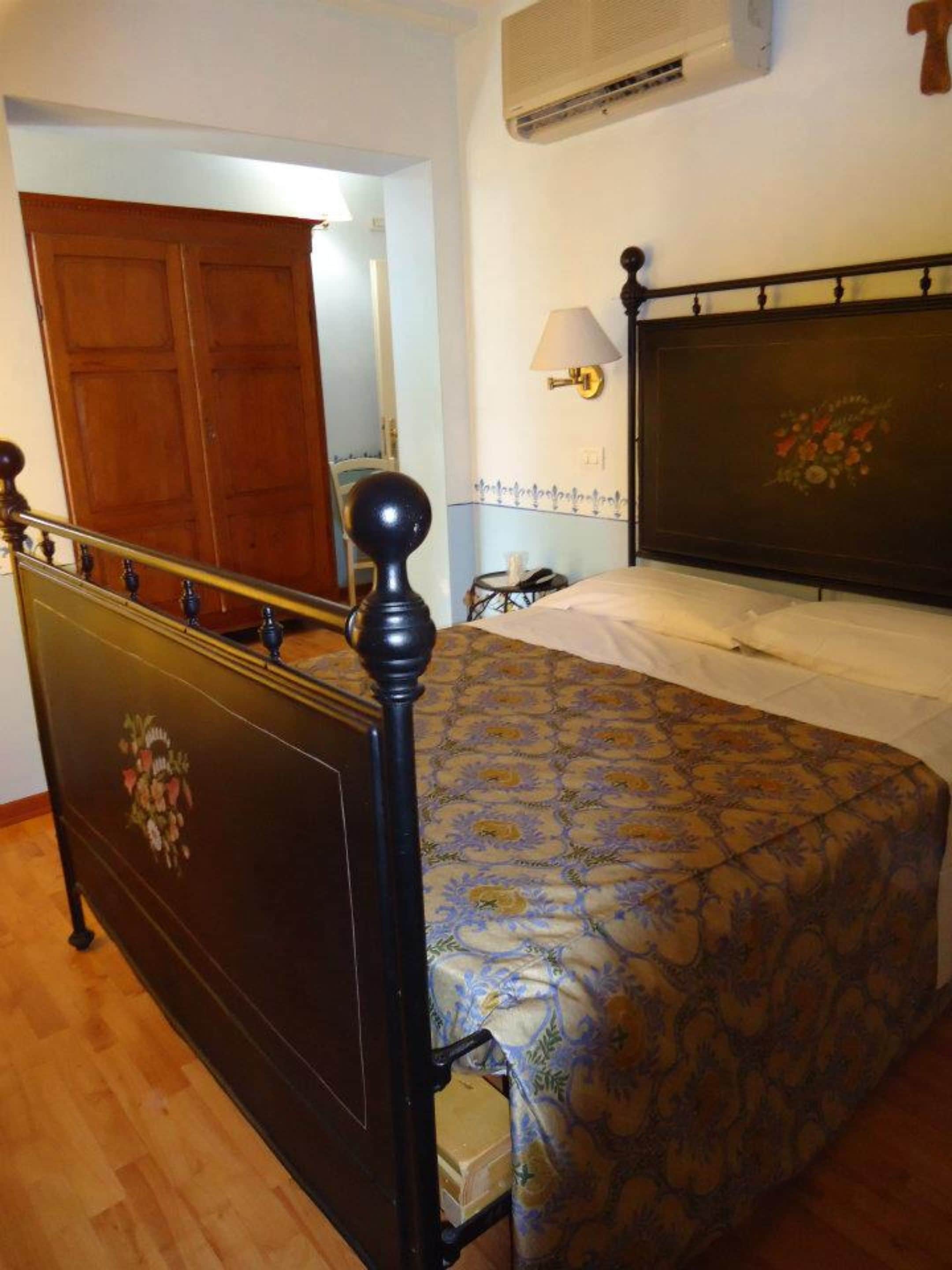 A bedroom with a wooden bed frame, a floral-patterned bedspread, a bedside lamp, and a wooden wardrobe.