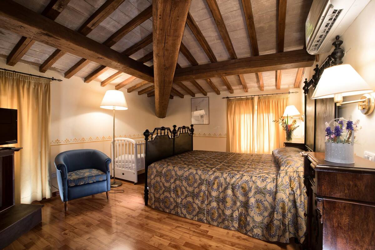 Un dormitorio con techo de madera, una cama con colcha estampada, un sillón azul, una mesita de noche con lámpara y un aparador.