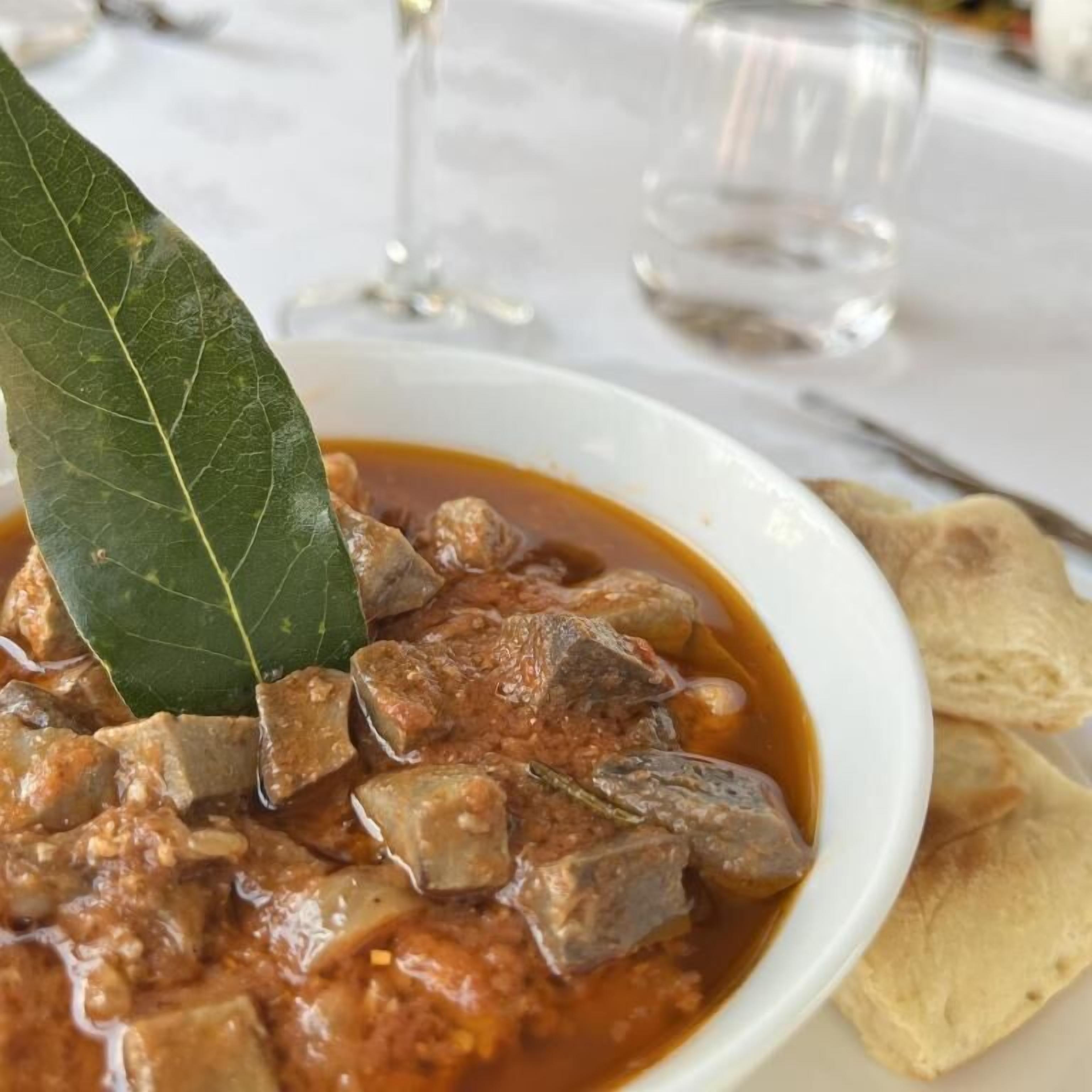 Un bol de ragoût contenant des morceaux de viande et garni d’une feuille de laurier, servi avec du pain plat à côté.