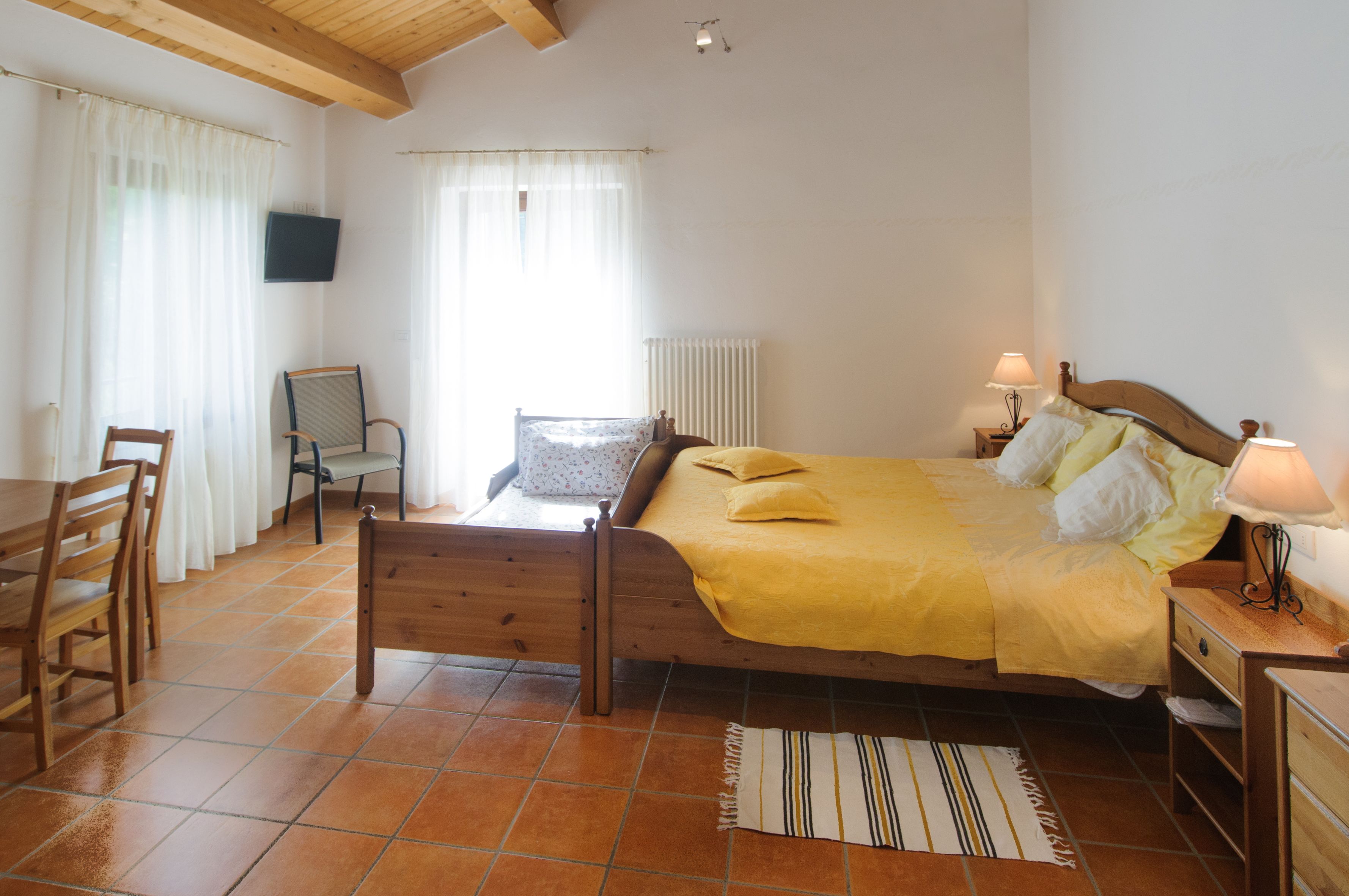 Une chambre à coucher avec un lit en bois, une chaise, une petite table, un t éléviseur fixé au mur et un radiateur.