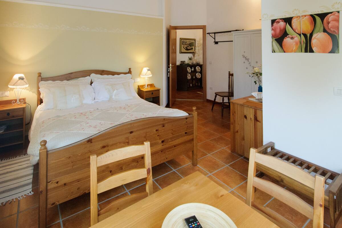 A wooden bed with white bedding, two bedside tables with lamps, a wooden chair, a small table with a remote control, and a painting of tulips on the wall.