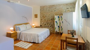 A bedroom with a stone wall, a bed with a white bedspread, a wooden nightstand, a bedside table with a lamp, a wooden chair, and a TV mounted on the wall. - B&B La Fenice (Mondaino)