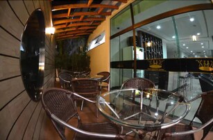 Un espacio de estar al aire libre con sillas de mimbre y una mesa de vidrio, con vistas a un restaurante con pared de cristal e iluminación interior.