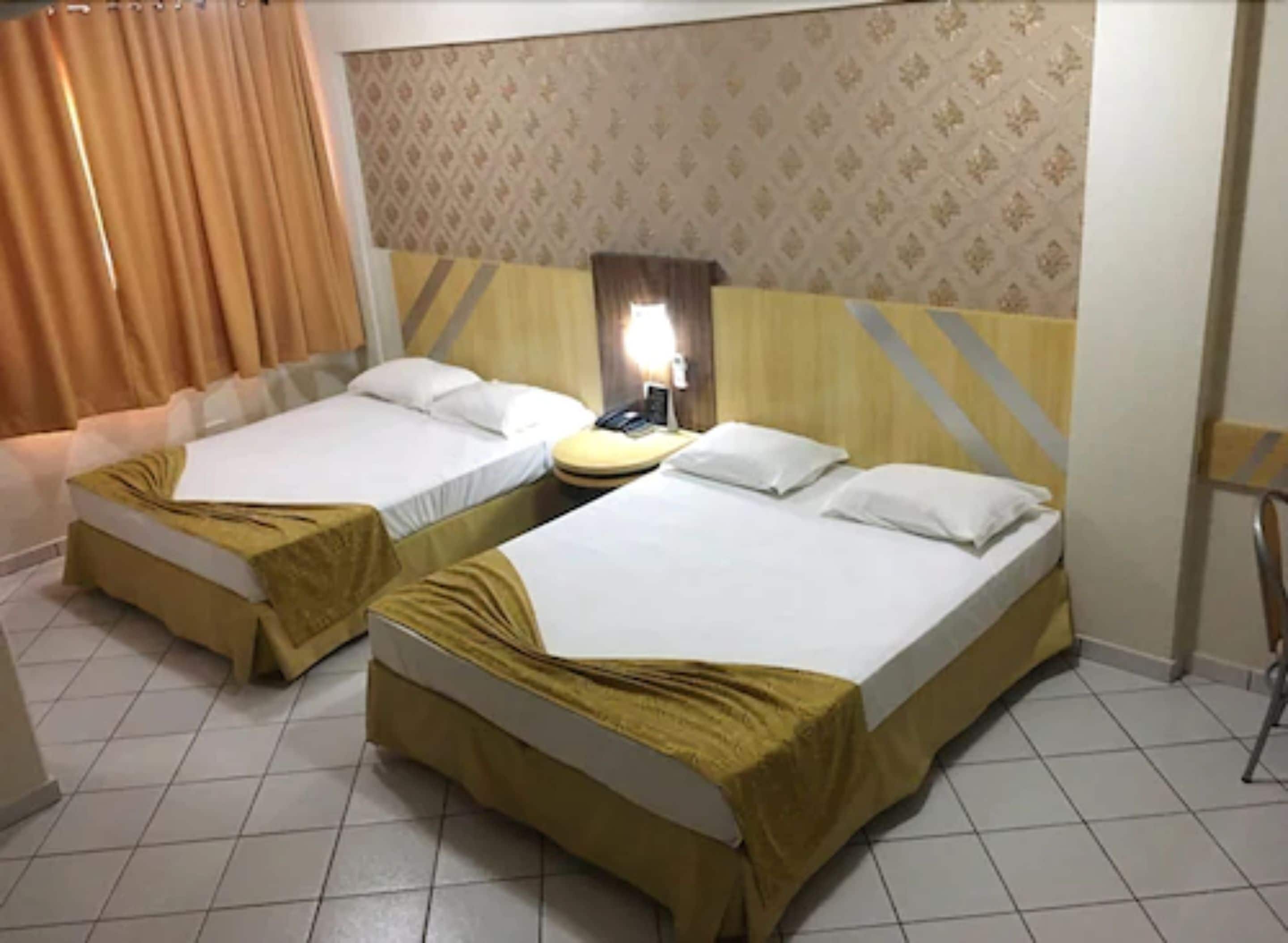 A hotel room with two beds, a bedside table with a lamp, and a patterned wallpaper.