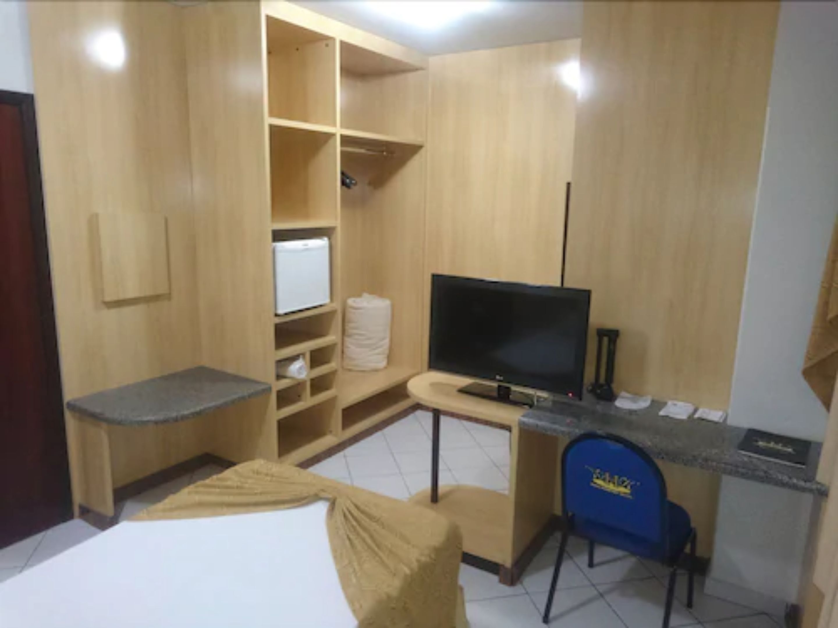 Quarto compacto com uma cama, escrivaninha com computador, televisão e guarda-roupa.